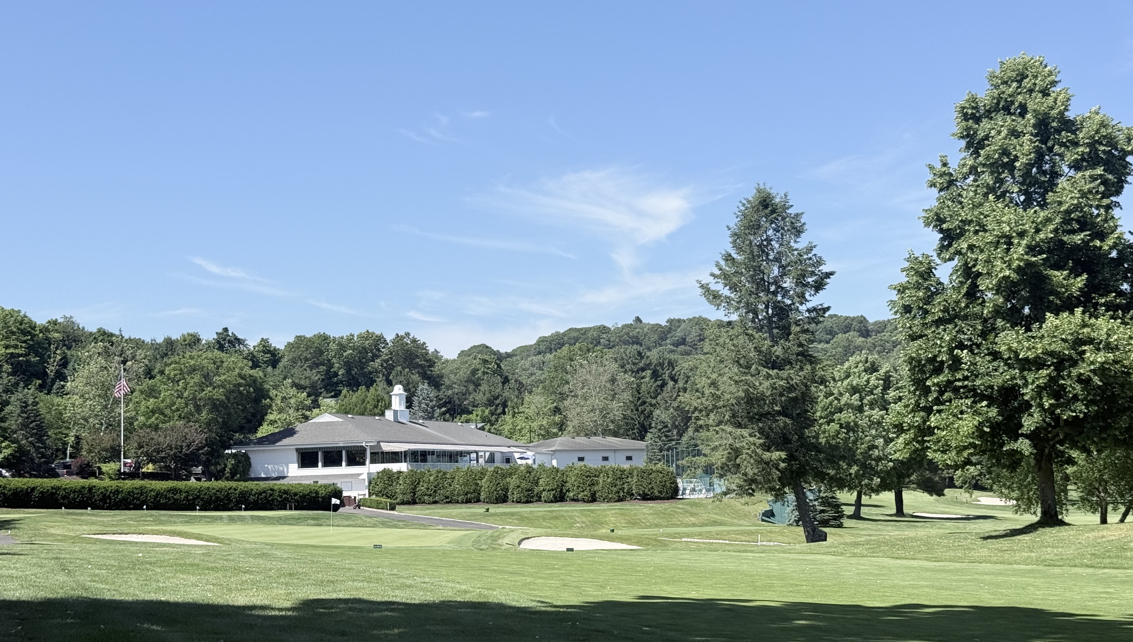 Brownson Country Club's white clubhouse with cupola and American flag overlooking manicured fairways, sand bunkers, and a putting green framed by tall evergreens in Huntington, Connecticut