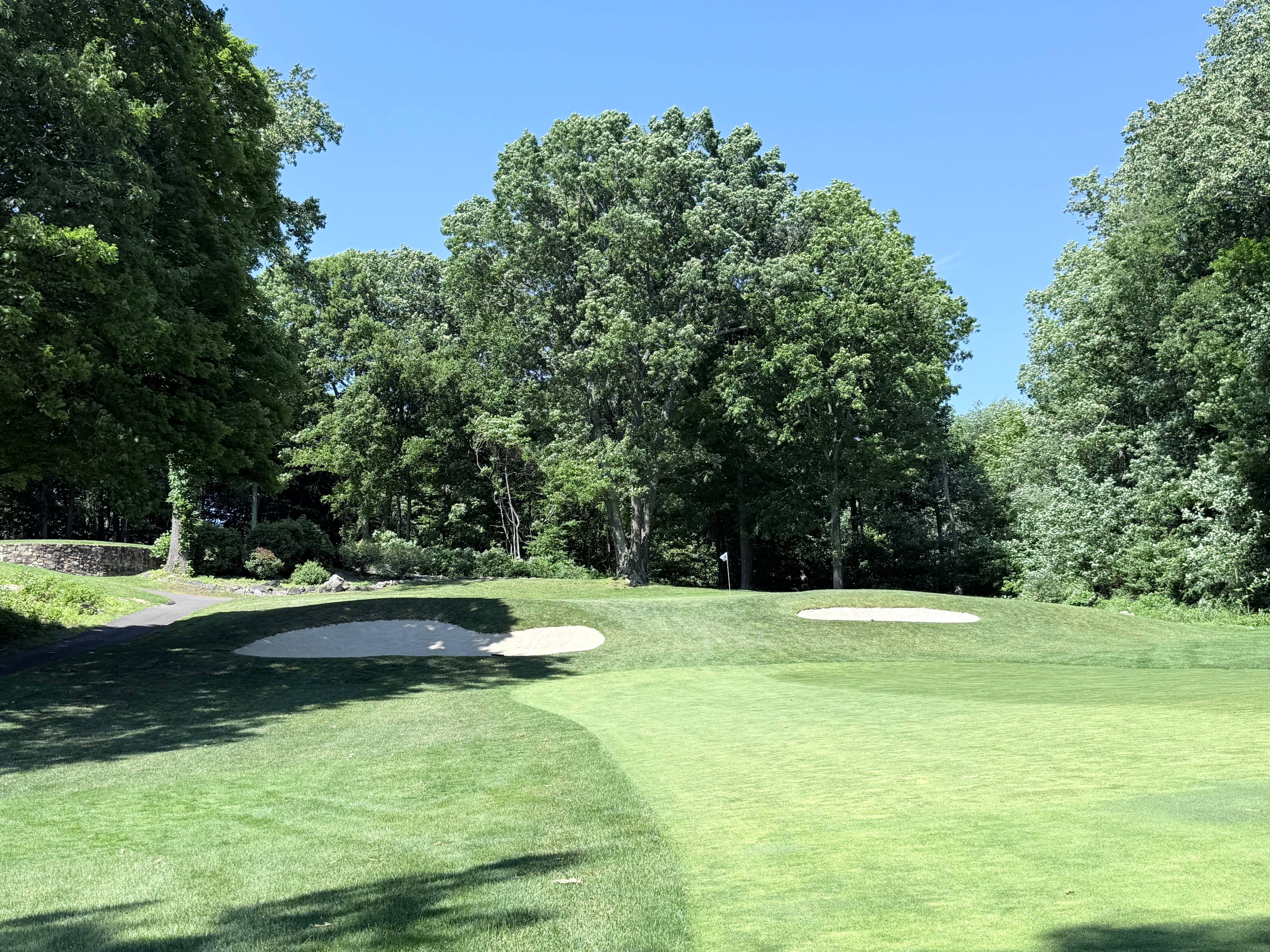 The 13th hole at Brownson Country Club, a manicured green flanked by white sand bunkers and framed by mature trees under a clear blue sky