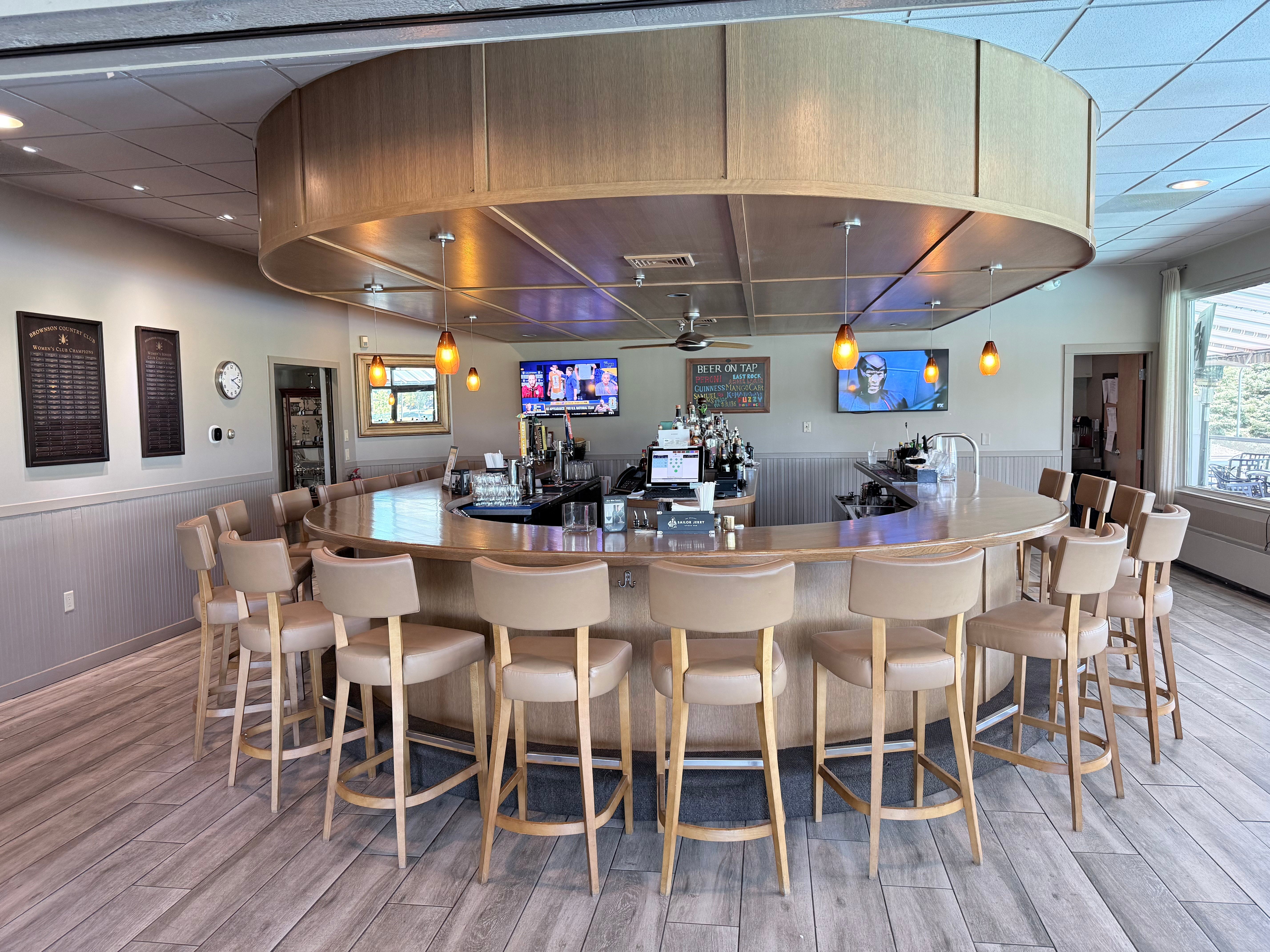 The Grille Bar at Brownson Country Club, a large oval wooden bar with cushioned leather stools, multiple TVs, a beer-on-tap chalkboard, and championship plaques on the walls