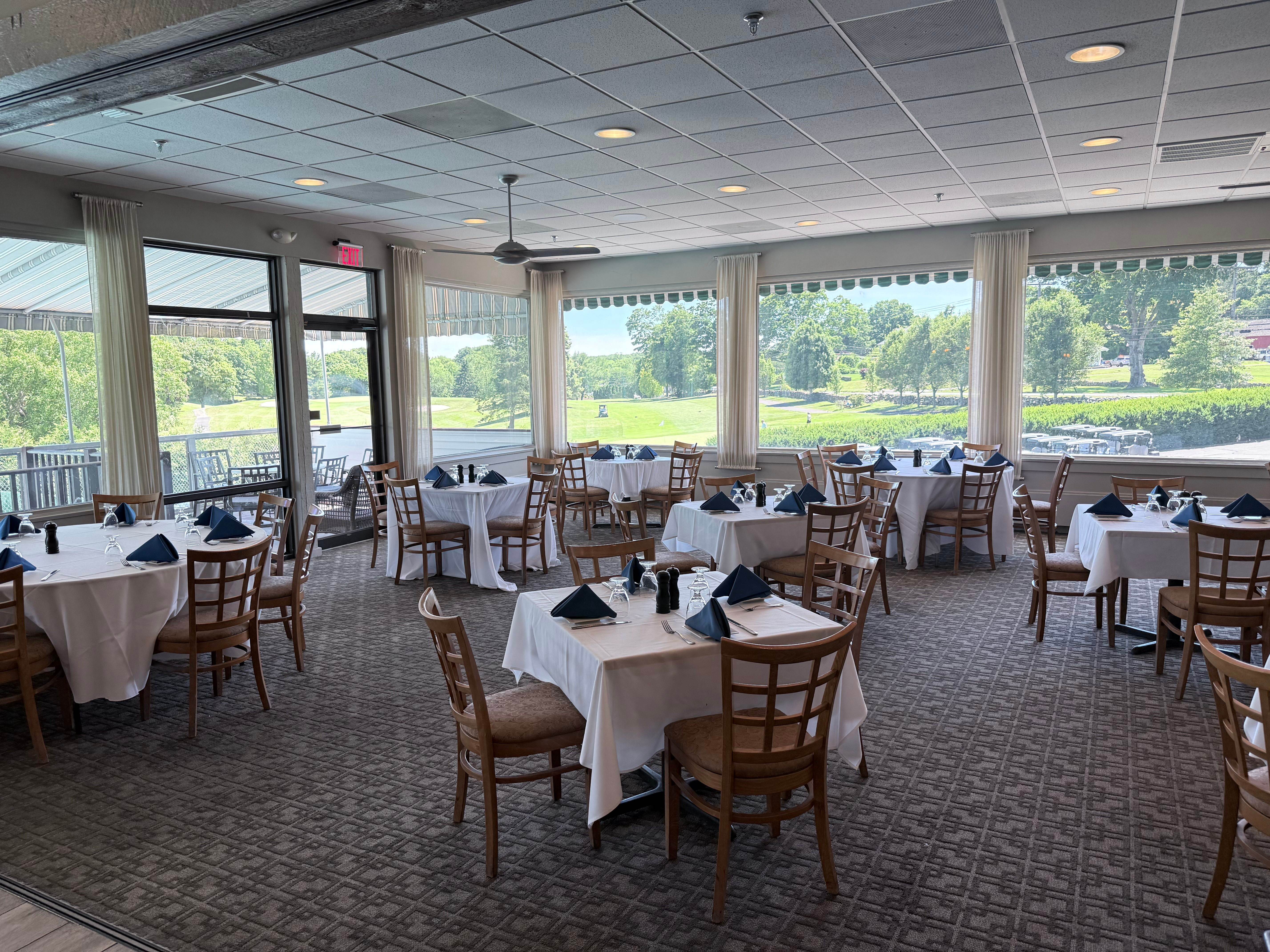 The Grille Room at Brownson Country Club with white-linen tables, navy napkins, and wooden chairs beside large windows overlooking the golf course
