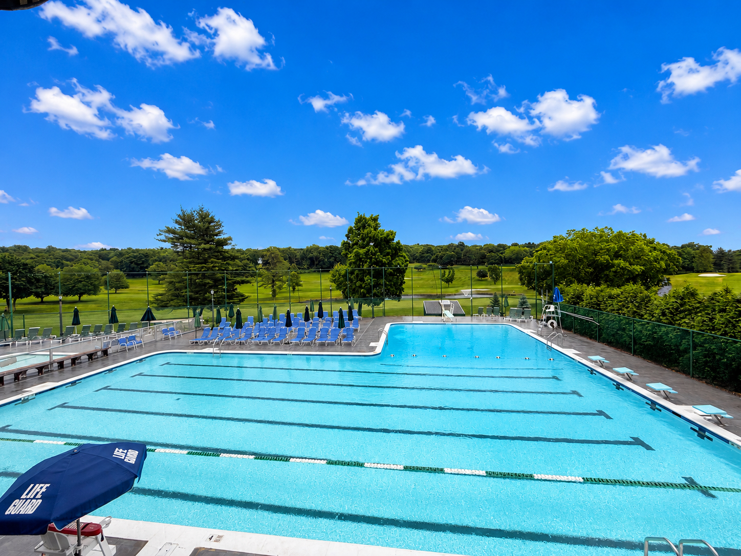 The Brownson Country Club outdoor pool complex with a multi-lane lap pool, diving boards, blue lounge chairs and umbrellas, overlooking the golf course