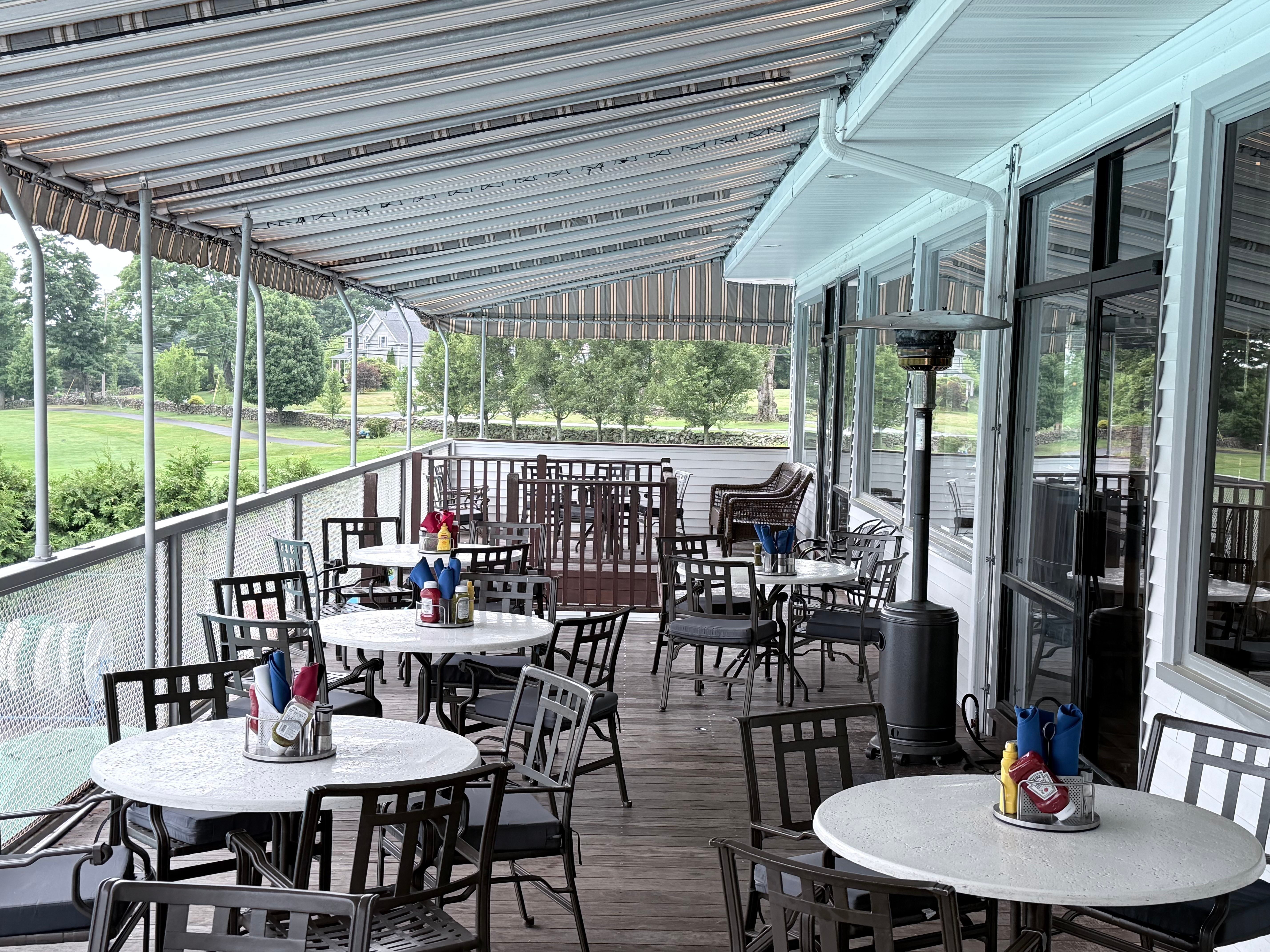 The covered Pool Deck at Brownson Country Club with round white bistro tables, patio chairs, a striped awning, and views of the golf course through the railing